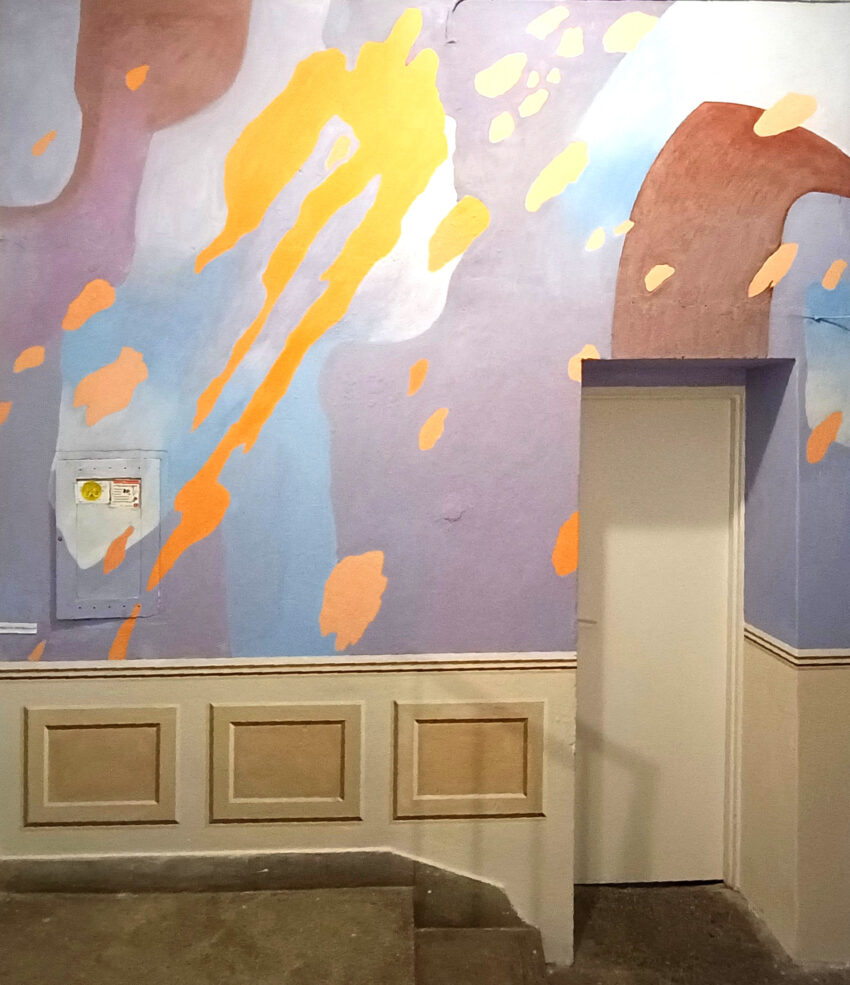 The photo shows a staircase leading to a closed white door, surrounded by a colourful abstract wall painting. The painting features pastel shades like lavender, blue, and rusty, along with bold orange streaks and patches. The lower part of the wall has a trompe-l'œil painting of classic panelling.