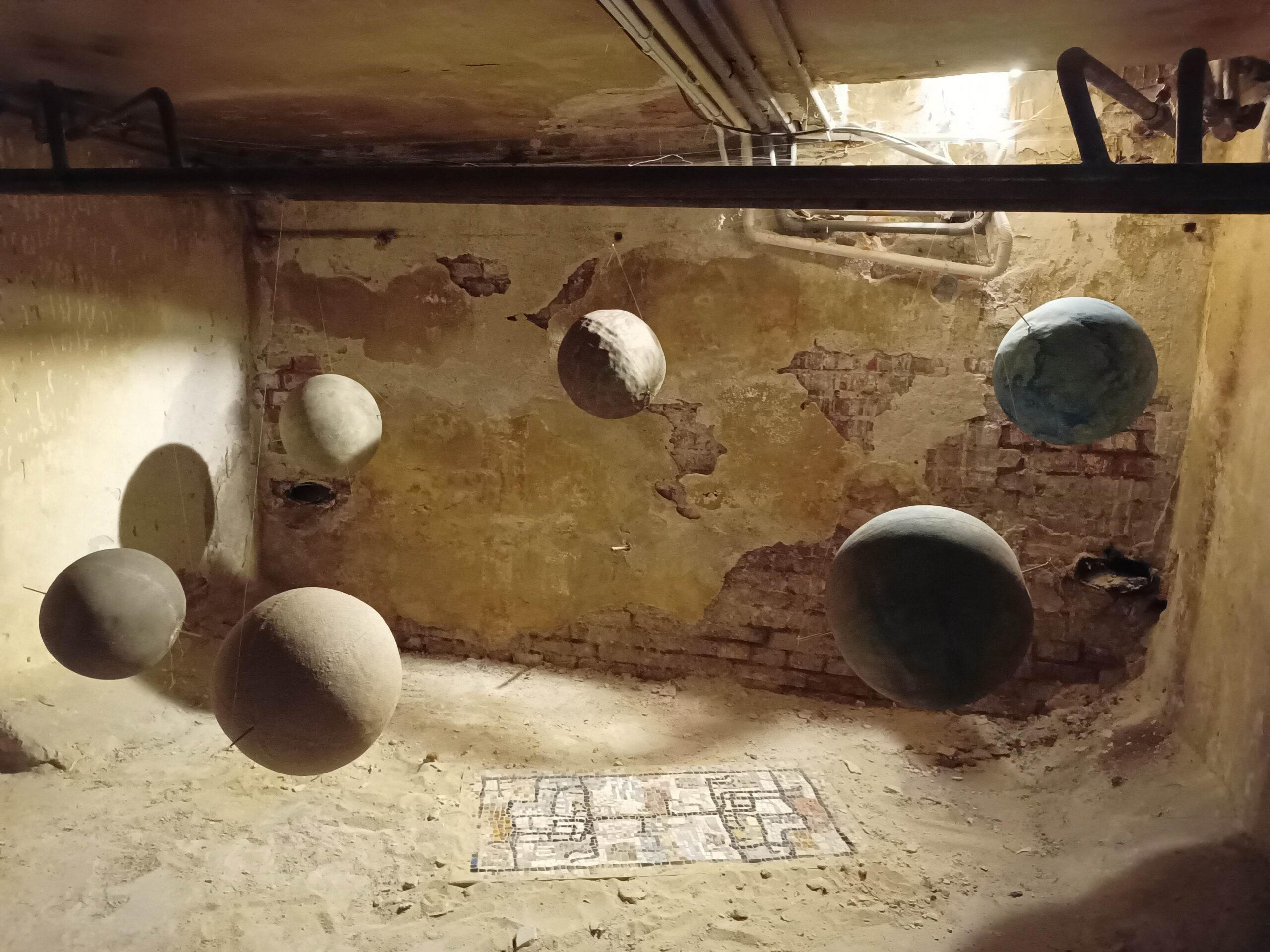 A photograph of an underground interior a with a gallery display of six linen globes floating above ground around a mosaic that is somewhat buried in sand on the ground.