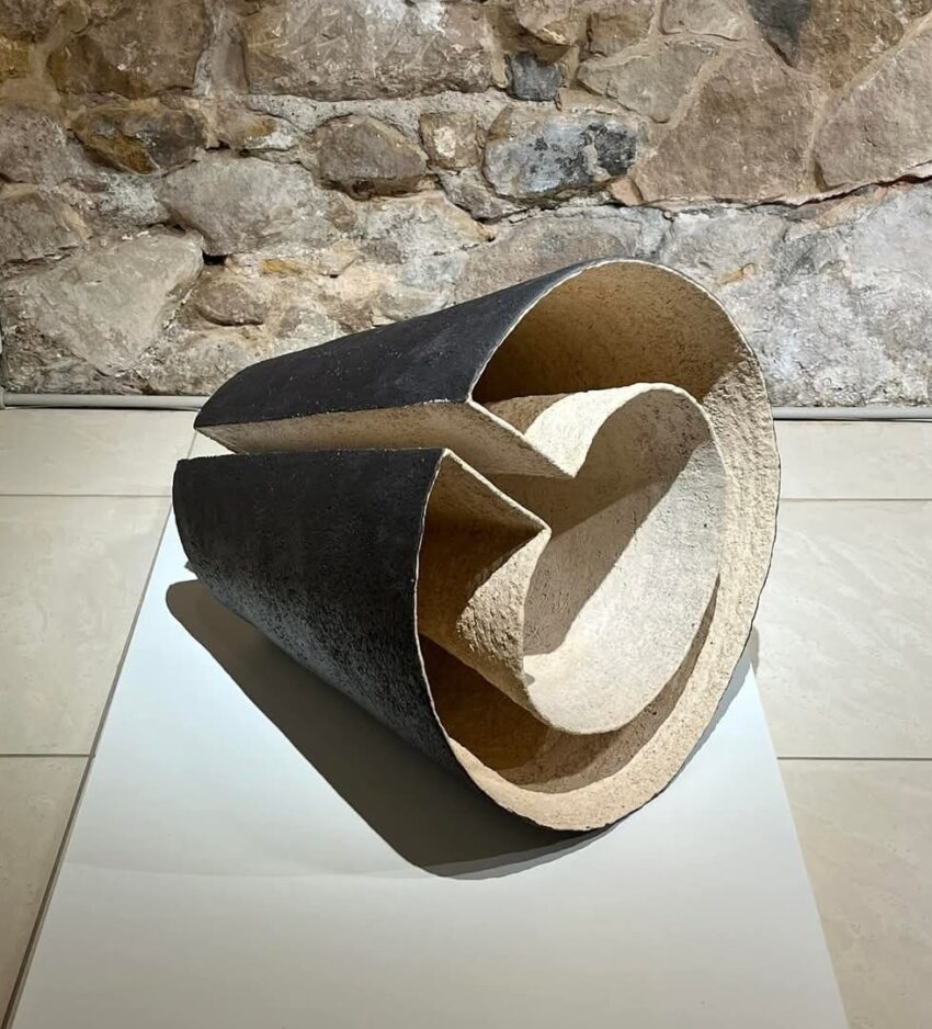 This ceramic sculpture features a hollow, cylindrical form with a dark, textured exterior and a smooth, beige interior. Partially sliced open, it reveals layered geometric cuts. Displayed on a white platform, the piece casts a shadow under gallery lighting. Made from chamotte clay and glaze, it was fired at 1100°C.