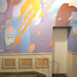 The photo shows a staircase leading to a closed white door, surrounded by a colourful abstract wall painting. The painting features pastel shades like lavender, blue, and rusty, along with bold orange streaks and patches. The lower part of the wall has a trompe-l'œil painting of classic panelling.