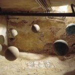 A photograph of an underground interior a with a gallery display of six linen globes floating above ground around a mosaic that is somewhat buried in sand on the ground.